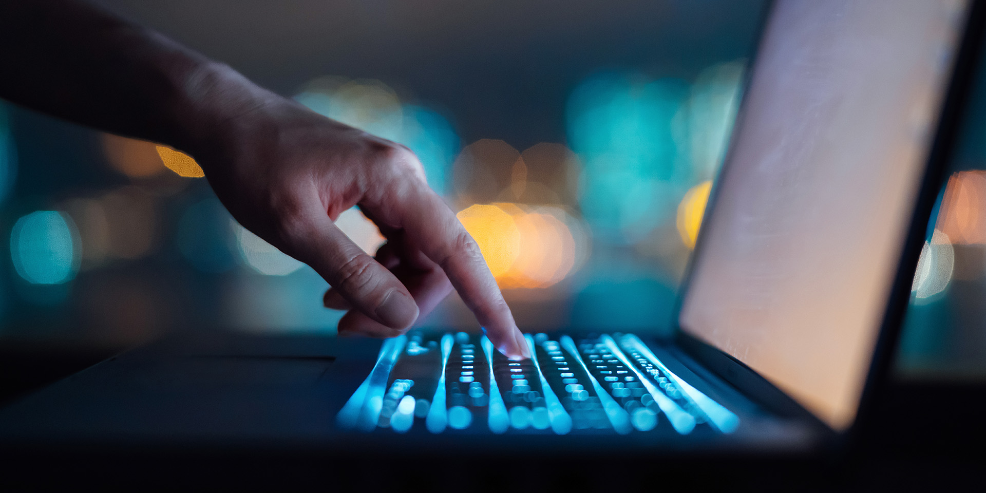 Close up of woman's hand typing on computer keyboard in the dark against colourful bokeh in background, working late on laptop at home