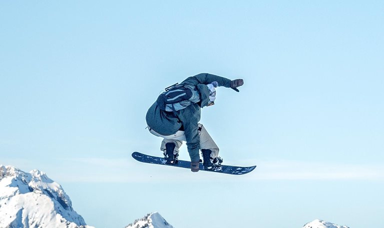 A snowboarder is captured midair performing a jump against a clear blue sky, with rugged, snow-covered mountains stretching across the background. The scene conveys motion and adventure, highlighting the contrast between the airborne rider and the vast alpine landscape below.