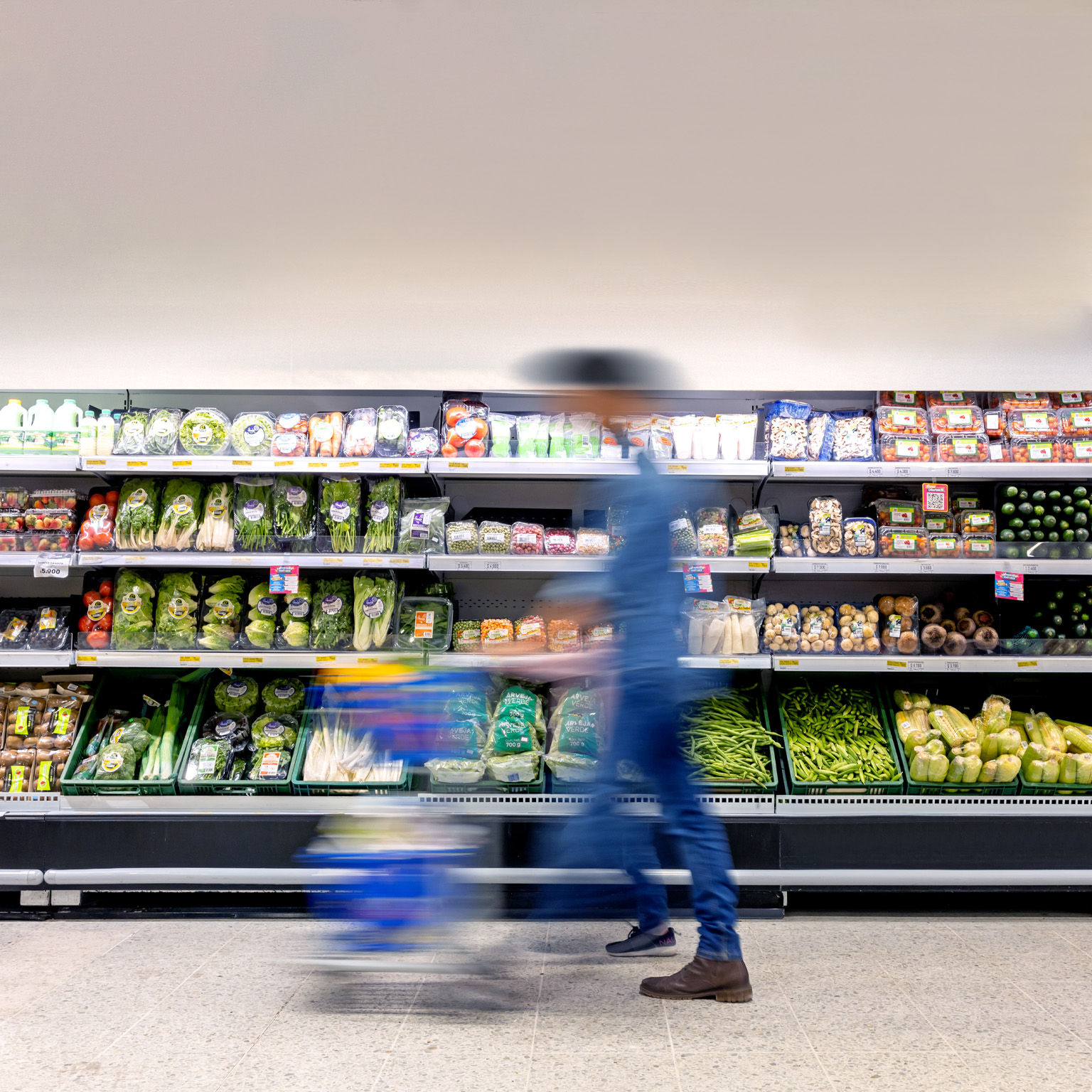 A man maneuvering a shopping cart through the grocery store, passing by the refrigerated produce section.