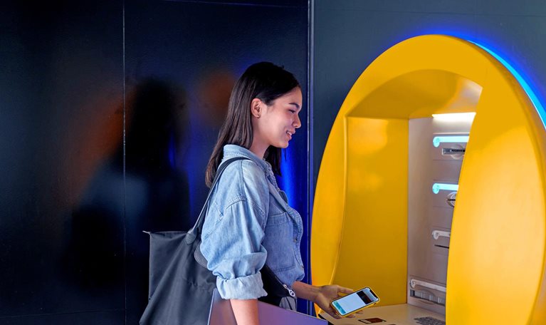 A woman with a smile on her face is standing at an ATM, holding a cell phone and laptop, as she prepares to withdraw money.
