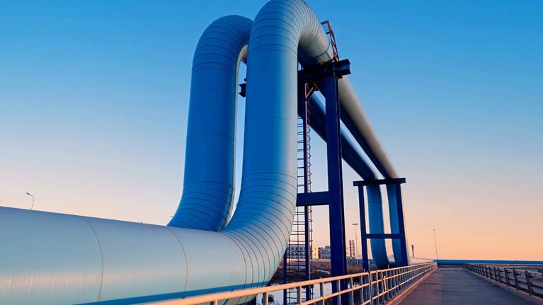 A large industrial pipeline rises and bends over a bridge-like structure, set against a clear blue sky at dusk. A paved walkway with railings runs alongside the pipes, leading the eye toward the horizon and giving the scene a clean, modern, and slightly futuristic feel.
