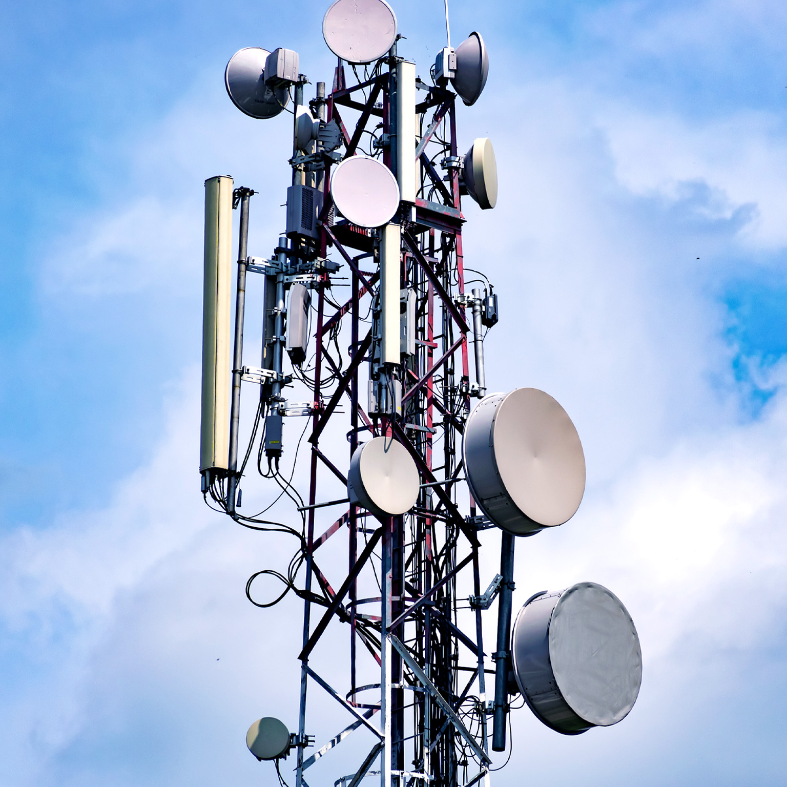 A tall telecommunications tower rises against a partly cloudy blue sky, covered with multiple circular and rectangular antennas.