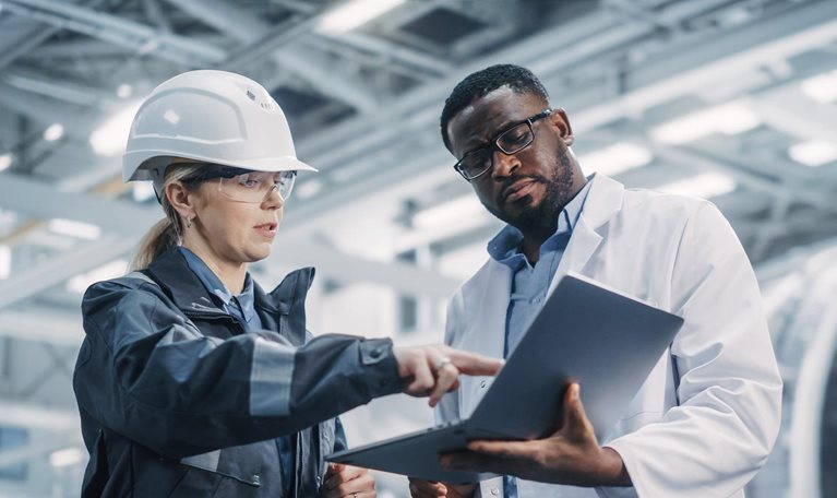 Team of Diverse Professional Heavy Industry Engineers Wearing Safety Uniform and Hard Hat Working on Laptop Computer. African American Technician and Female Worker Talking on a Meeting in a Factory. - stock photo