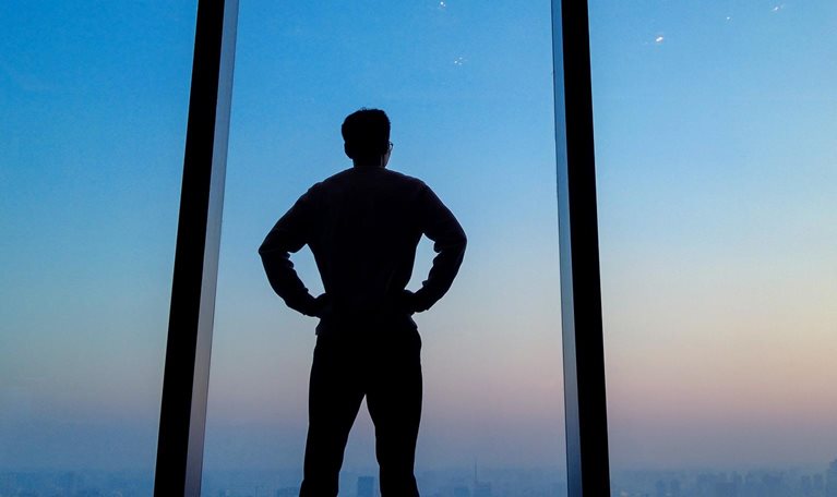 Young man staring out floor to ceiling office window looking over city skyline