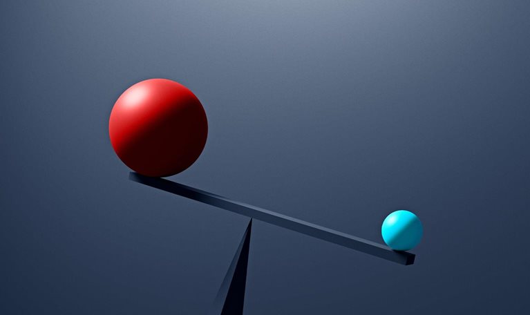 A large red sphere and a much smaller, light-blue sphere rest on opposite ends of a simple seesaw, which is unbalanced with the smaller blue sphere outweighing the red one. The scene is set against a dark, neutral background, emphasizing the contrast in size and weight between the two spheres.