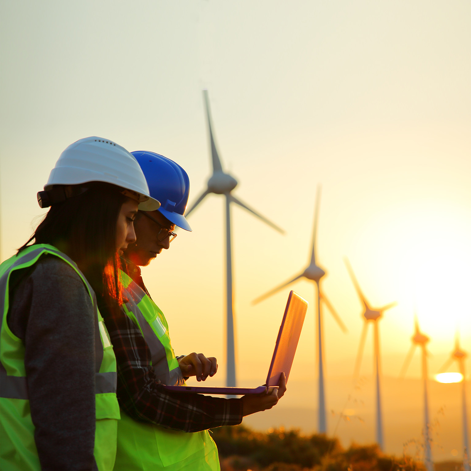 photo of two workers looking at laptop outside wind turbines with the backdrop of a beautiful sunset