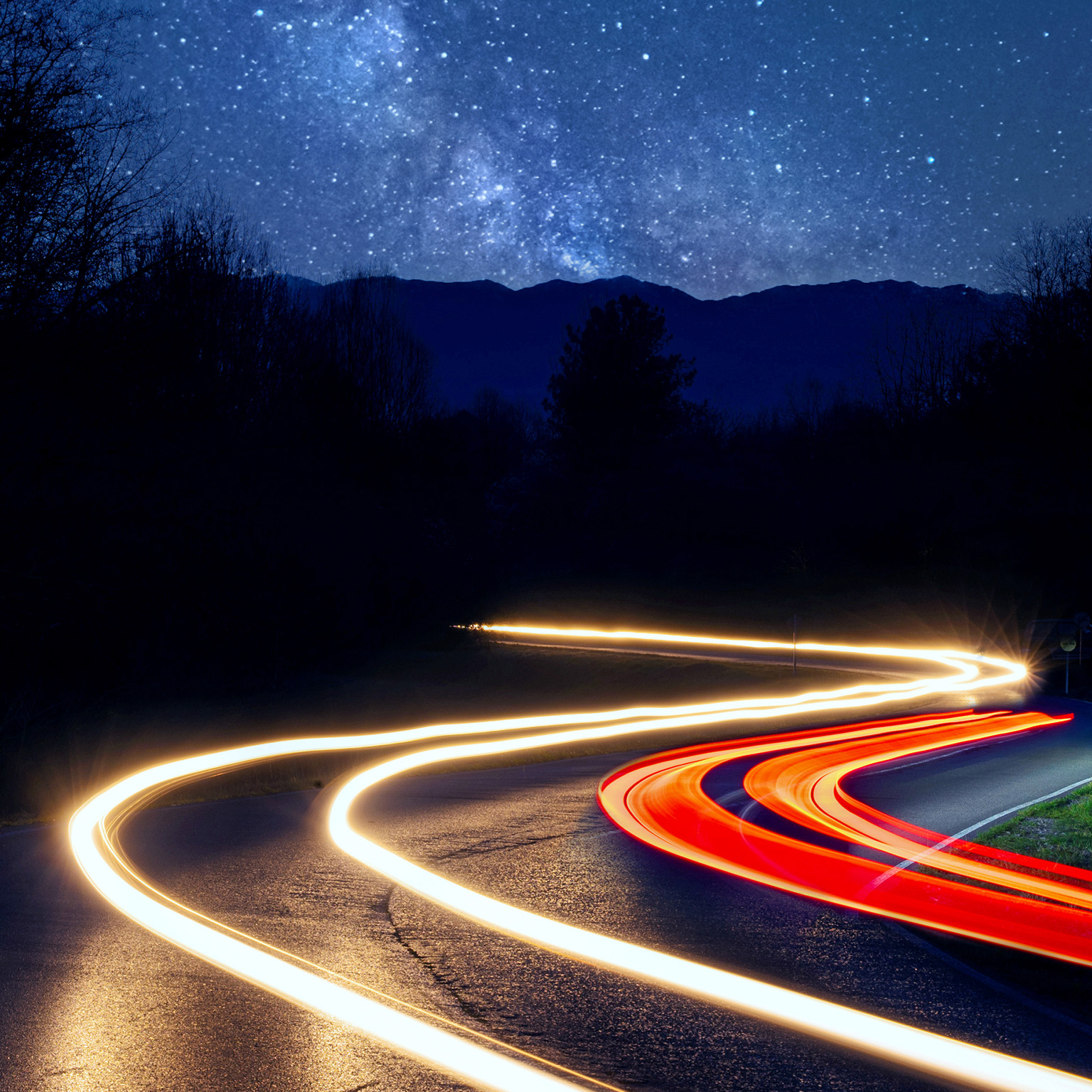car headlight trails driving on a road through a forest on a starry night with the milky way and mountains in the background