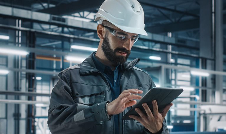 Portrait of Automotive Industry Engineer in Safety Glasses and Uniform Using Laptop at Car Factory Facility. Professional Assembly Plant Specialist Working on Manufacturing Modern Electric Vehicles.