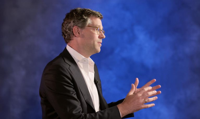 Erikhans Kok wearing glasses, a dark suit jacket, and a white shirt gestures with both hands while facing to the right against a blue background.