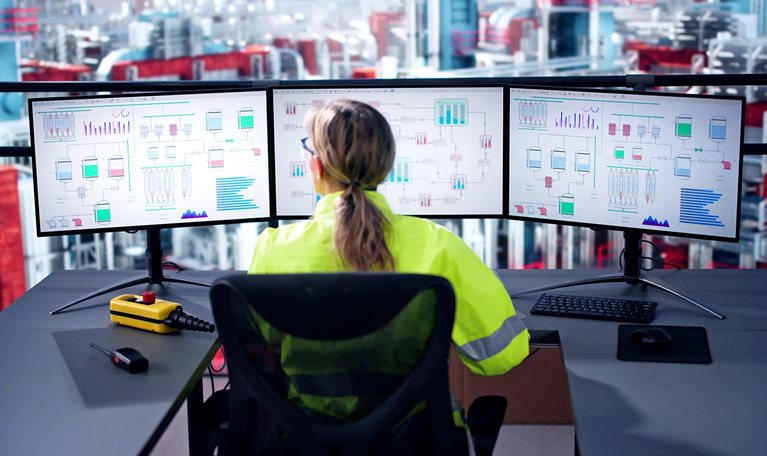 Image of an engineer operating an industrial data automation system using multiple computer screens with a factory in the background.