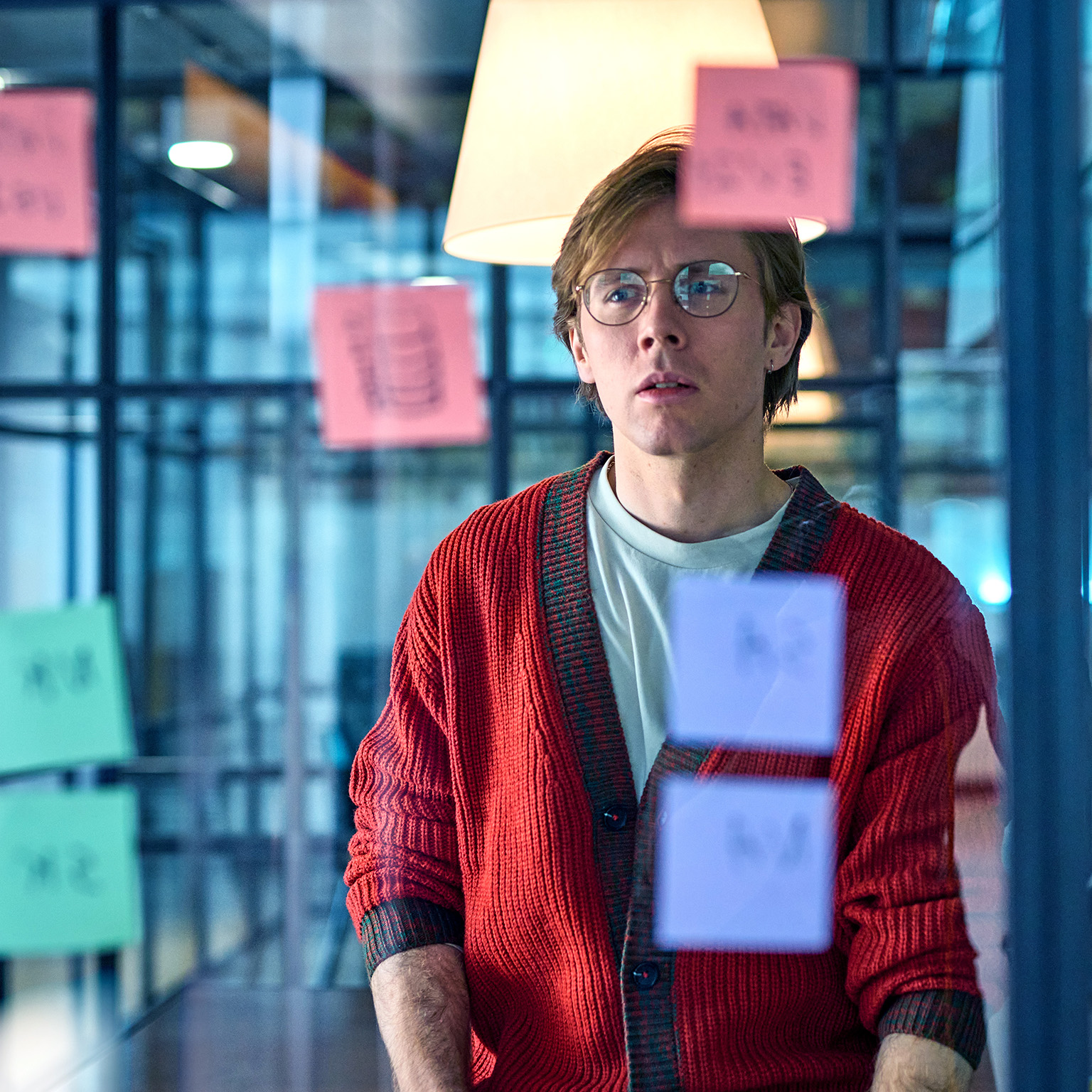 Man in his 30s wearing glasses reading post-it notes, curious expression, problem solving, planning, strategy