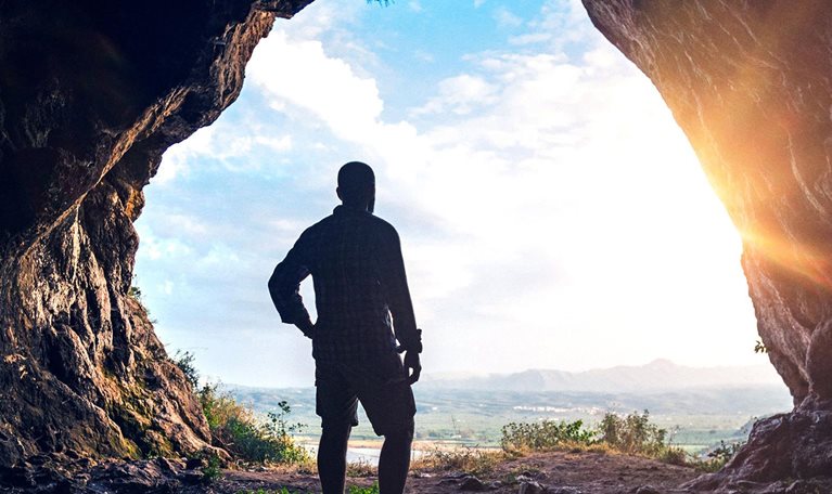 A hiker looks out over a sunset from inside the large opening of a cave