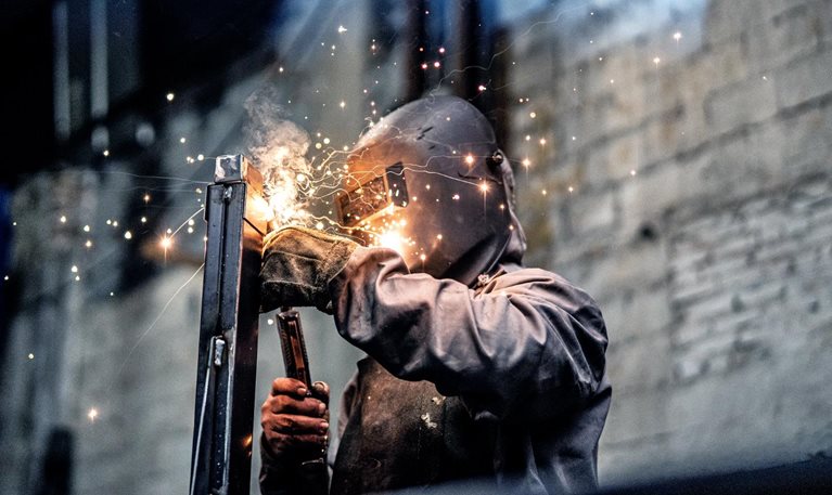An individual clad in protective welding attire, working on a steel component while sparks scatter.