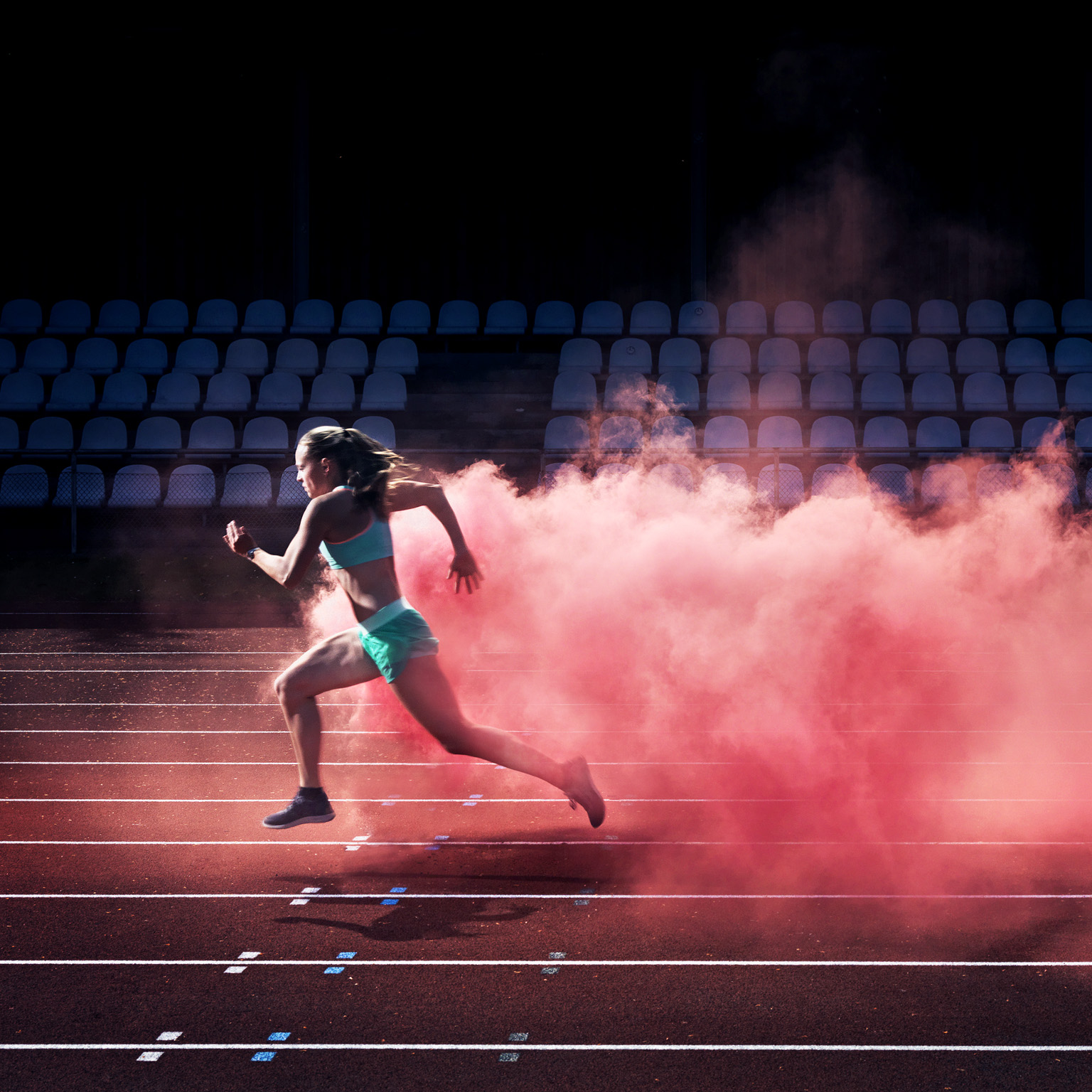 A determined female athlete races along a track, with a vibrant cloud of red smoke trailing behind her. 