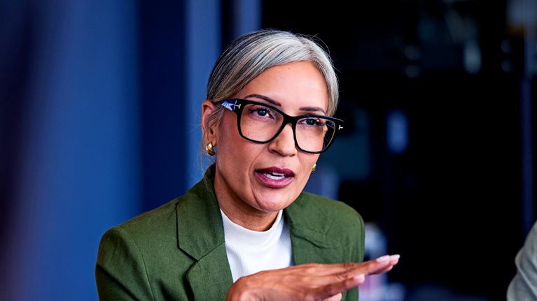 A mature businesswoman in a green blazer and glasses is speaking confidently while gesturing with her hands during a boardroom meeting.