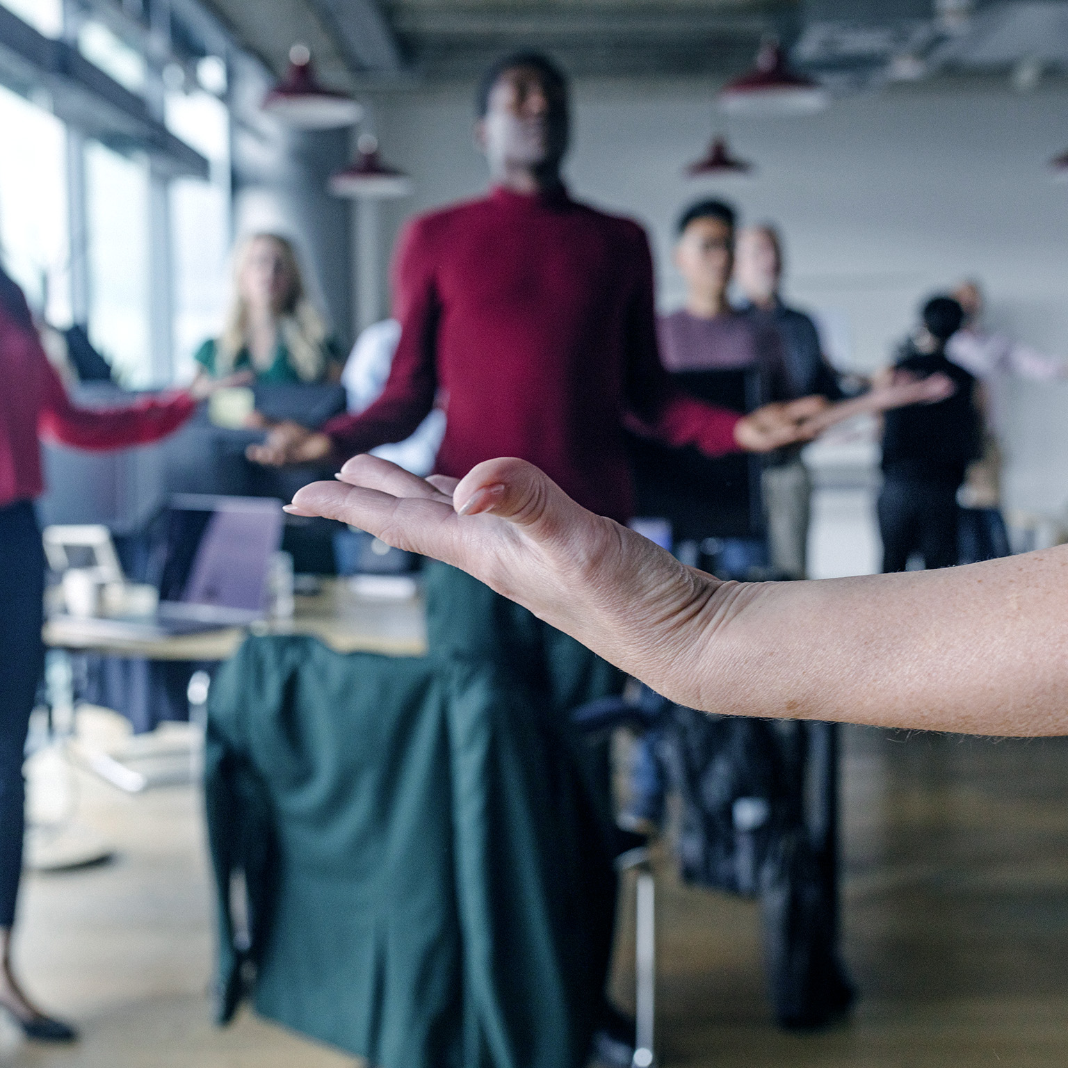 Close-up of a woman's upward facing hand in the foreground, with a blurred group of coworkers meditating in the background.