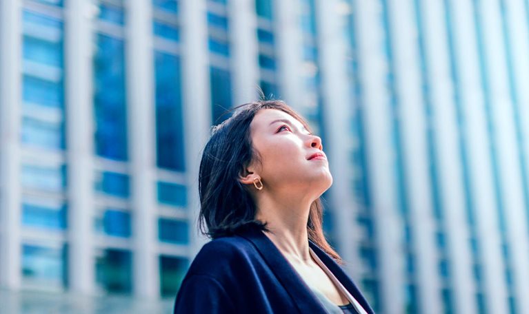 A modern business district with tall buildings is the background for a young, confident businesswoman looking up at the sky from a low angle.
