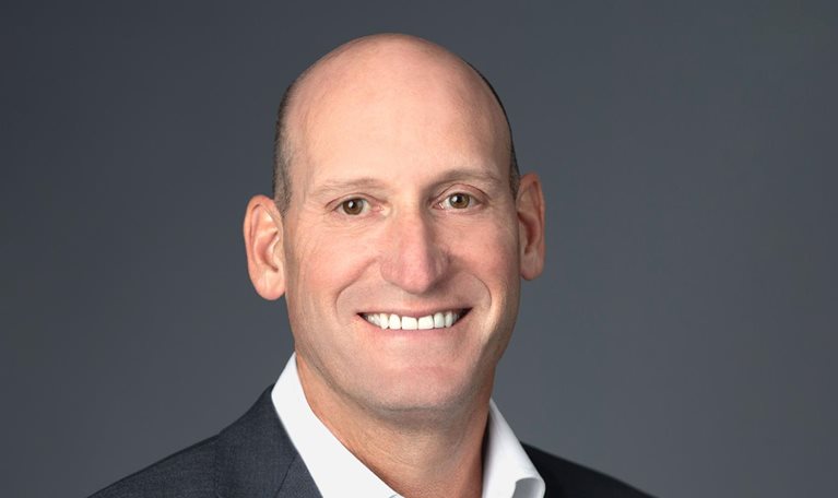 David Gitlin is standing against a neutral gray background, dressed in a dark suit jacket and white shirt, with a smile on his face.