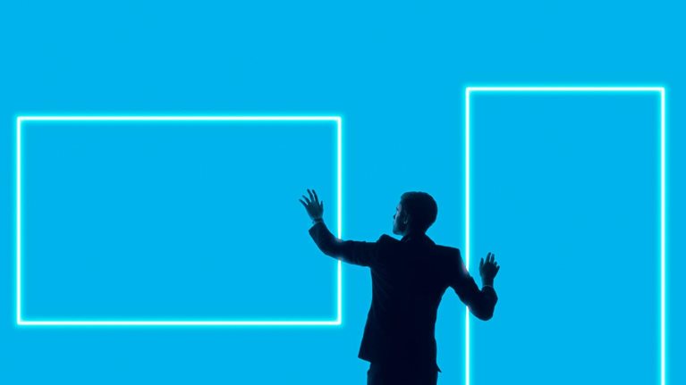 A silhouetted man in a suit stands against a bright blue background, reaching toward two glowing rectangular outlines that resemble digital screens or frames.