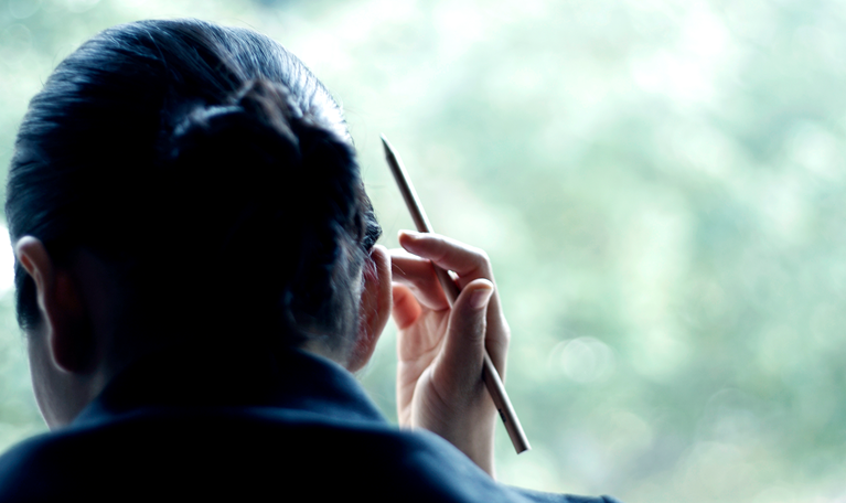 A person in a dark suit, seen from behind, holding a pen and looking out a window toward blurred green foliage.