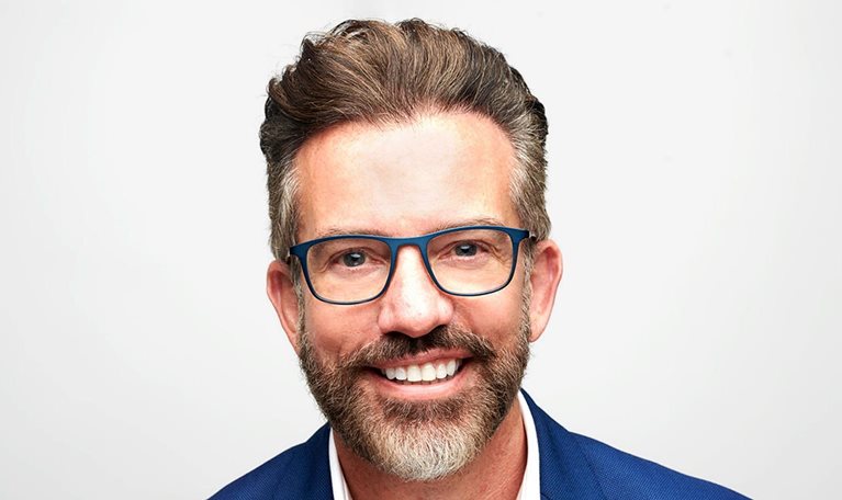 Nick Jeffery smiling warmly at the camera, wearing glasses and a blue suit jacket over a white shirt. He has neatly styled brown hair and a trimmed salt-and-pepper beard, set against a clean, light background.