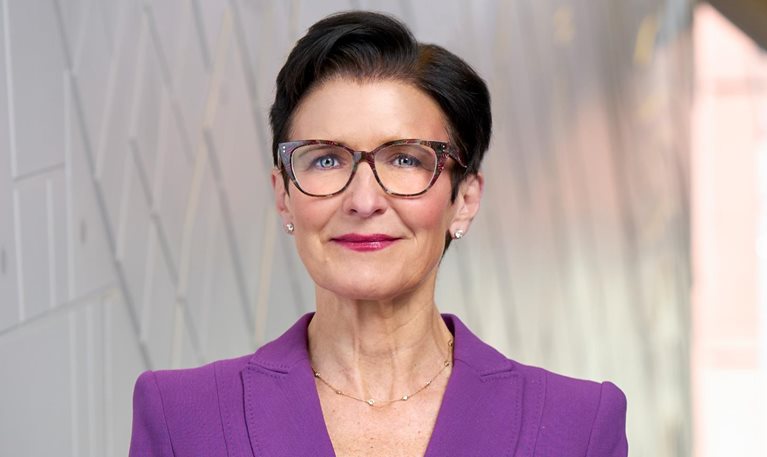 A portrait of Jane Fraser with short dark hair wearing glasses and a purple blazer over a black top. She stands against a softly blurred, modern interior background and looks directly at the camera with a confident, composed expression.