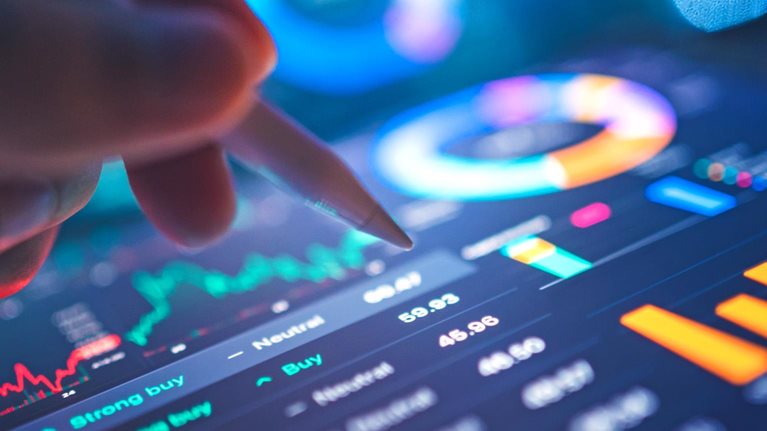 A hand uses a stylus to interact with a tablet displaying financial data and charts, including candlestick charts and pie charts. The screen glows with blue and orange hues.