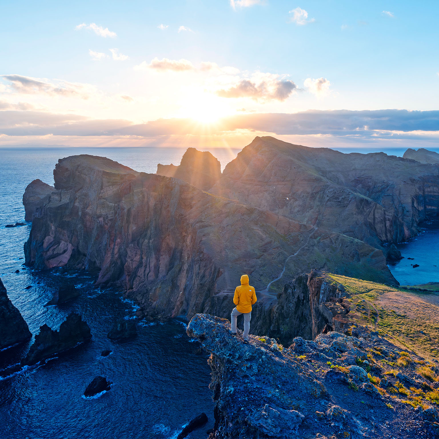  A person in a yellow jacket stands on a rocky cliff, looking out at the wide ocean and the sun rising over a mountainous coastline. 
