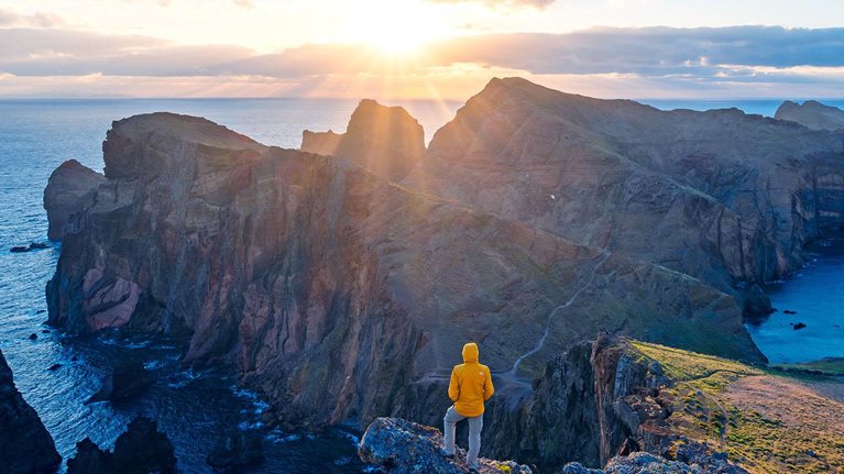 A person in a yellow jacket stands on a rocky cliff, looking out at the wide ocean and the sun rising over a mountainous coastline.