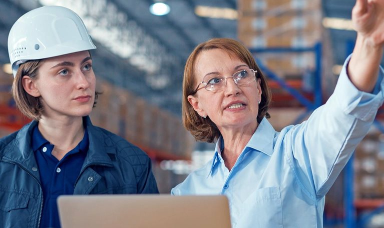 Two women in a warehouse reviewing information on a laptop and tablet, with one wearing a hard hat while the other gestures toward the facility.