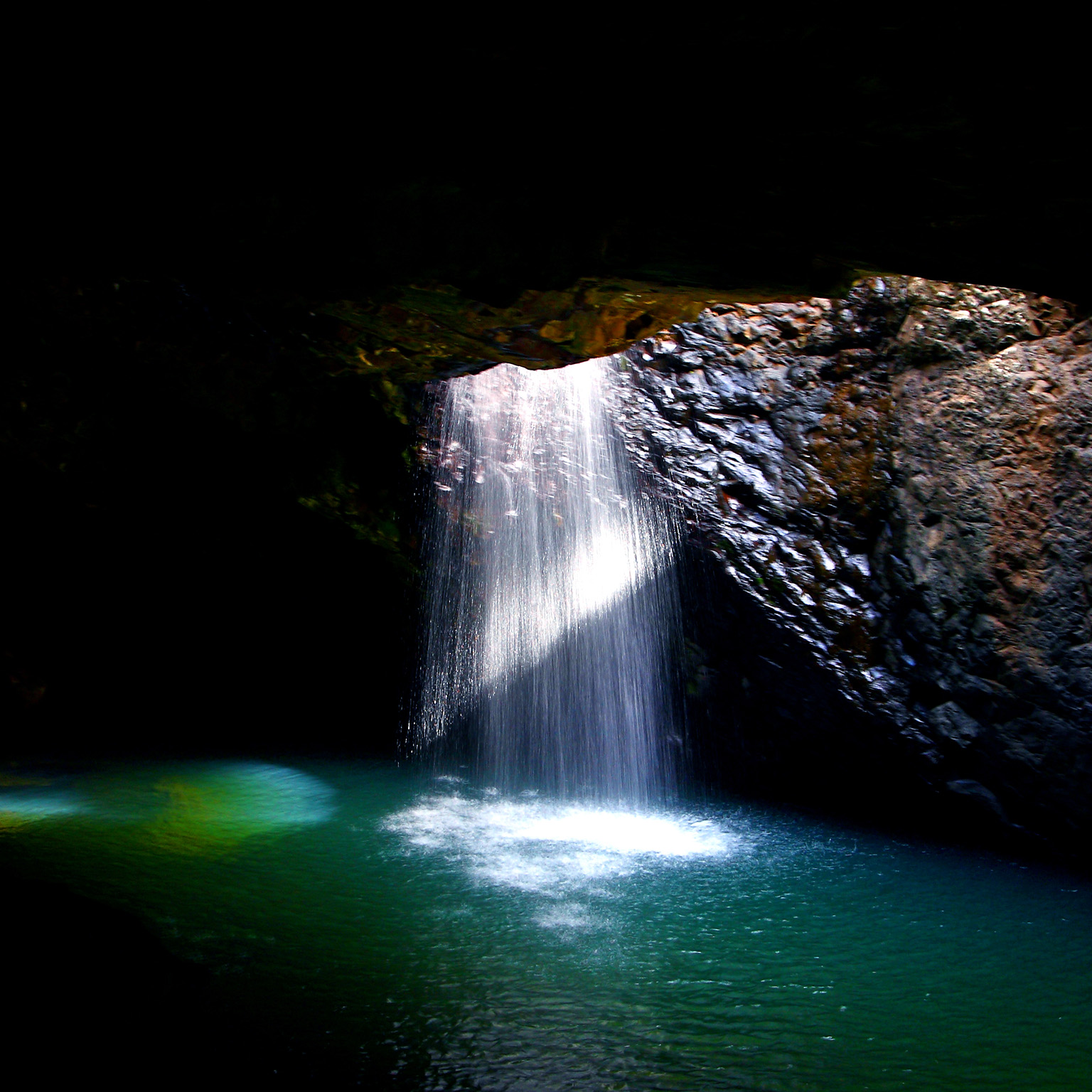 Sunlight streams through a cave opening, illuminating a stunning waterfall plunging into a vibrant turquoise pool below