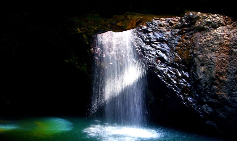 Sunlight streams through a cave opening, illuminating a stunning waterfall plunging into a vibrant turquoise pool below