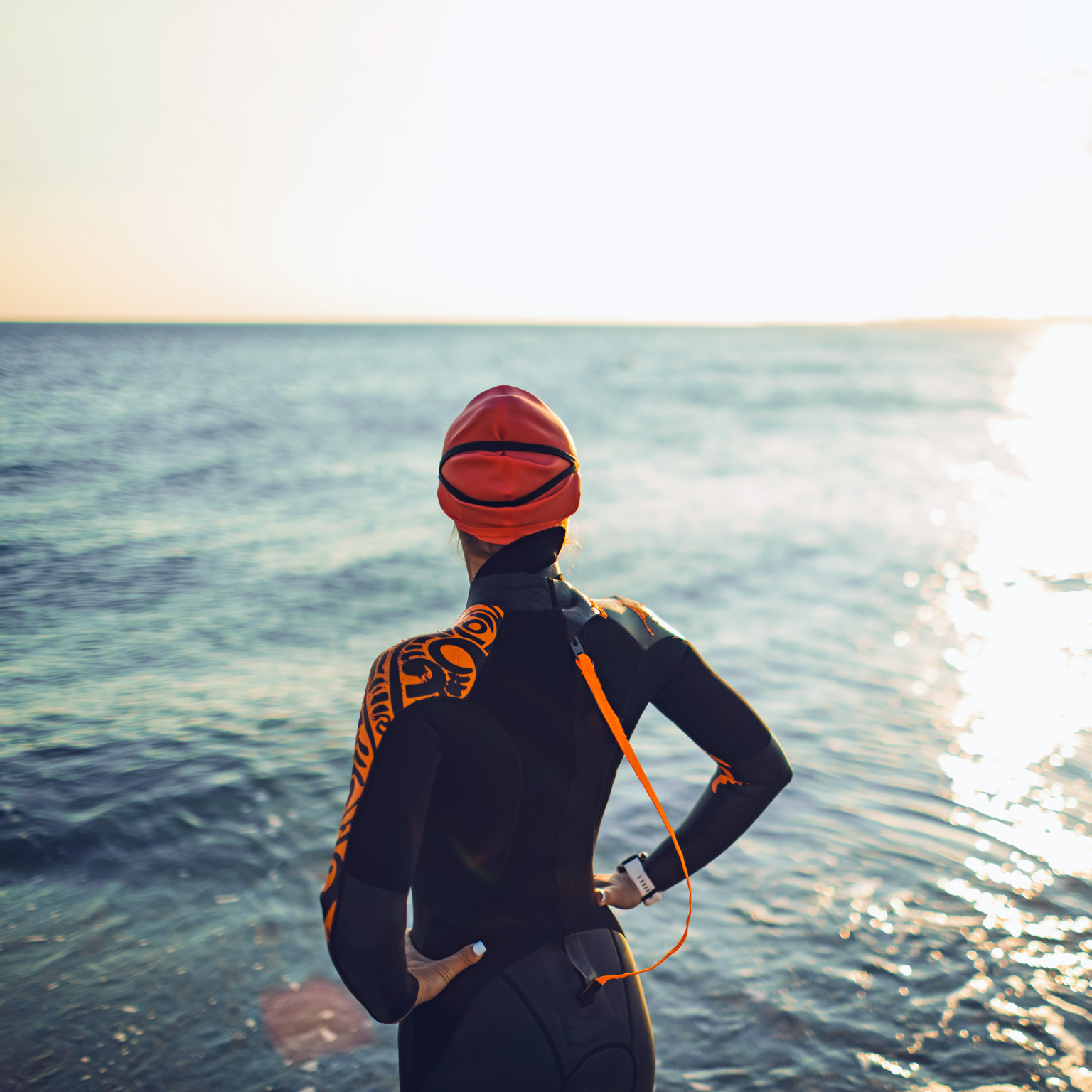 A triatalete wearing a wetsuit and swim cap stands facing a calm ocean. Her back is to the viewer as she takes in the setting sun. 