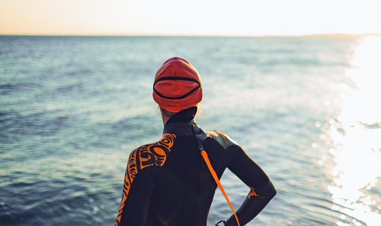 A triatalete wearing a wetsuit and swim cap stands facing a calm ocean. Her back is to the viewer as she takes in the setting sun.