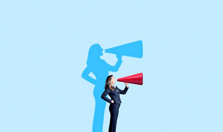 A woman in a business suit stands on a blue platform, speaking energetically into a red megaphone. Behind her, a blue shadow on the light-blue background mimics her pose, also holding a megaphone.