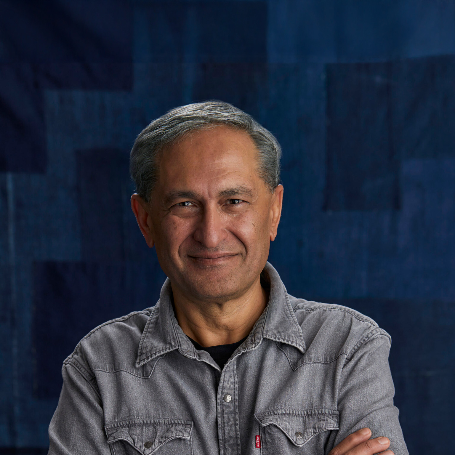 A portrait of a Harmit Singh with short hair and a slight smile, looking directly at the camera. He is wearing a gray button-up shirt and is set against a dark blue textured background.