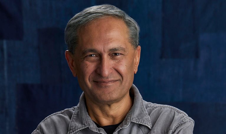 A portrait of a Harmit Singh with short hair and a slight smile, looking directly at the camera. He is wearing a gray button-up shirt and is set against a dark blue textured background.