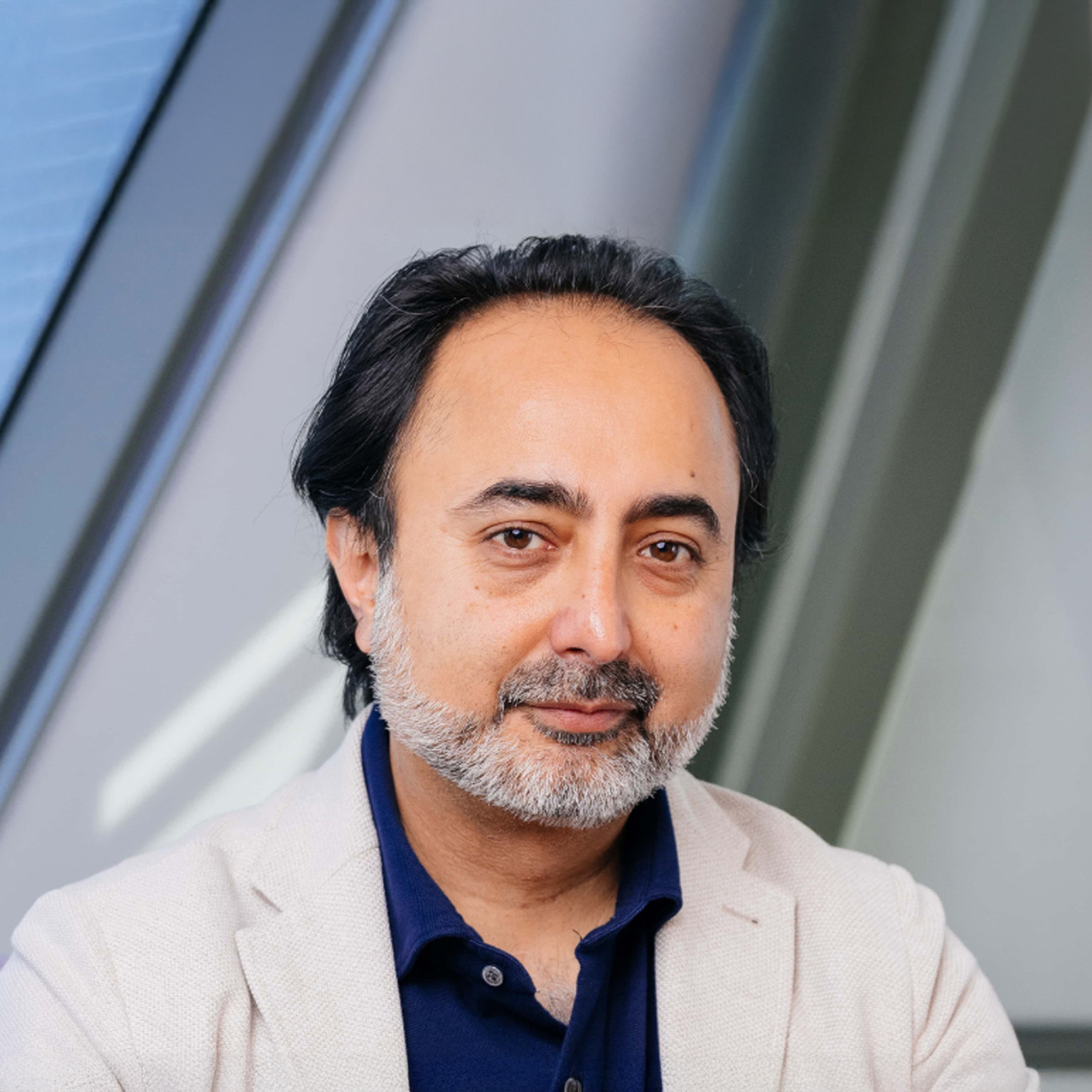 Hemant Taneja with dark hair and a neatly trimmed gray beard looking directly at the camera with a calm, neutral expression. He is wearing a light-colored blazer over a dark blue shirt, standing against a softly blurred indoor background with angled architectural lines.