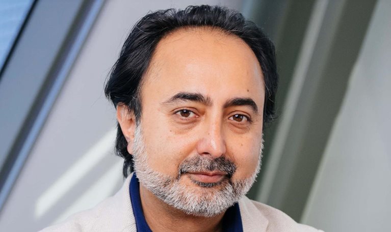 Hemant Taneja with dark hair and a neatly trimmed gray beard looking directly at the camera with a calm, neutral expression. He is wearing a light-colored blazer over a dark blue shirt, standing against a softly blurred indoor background with angled architectural lines.