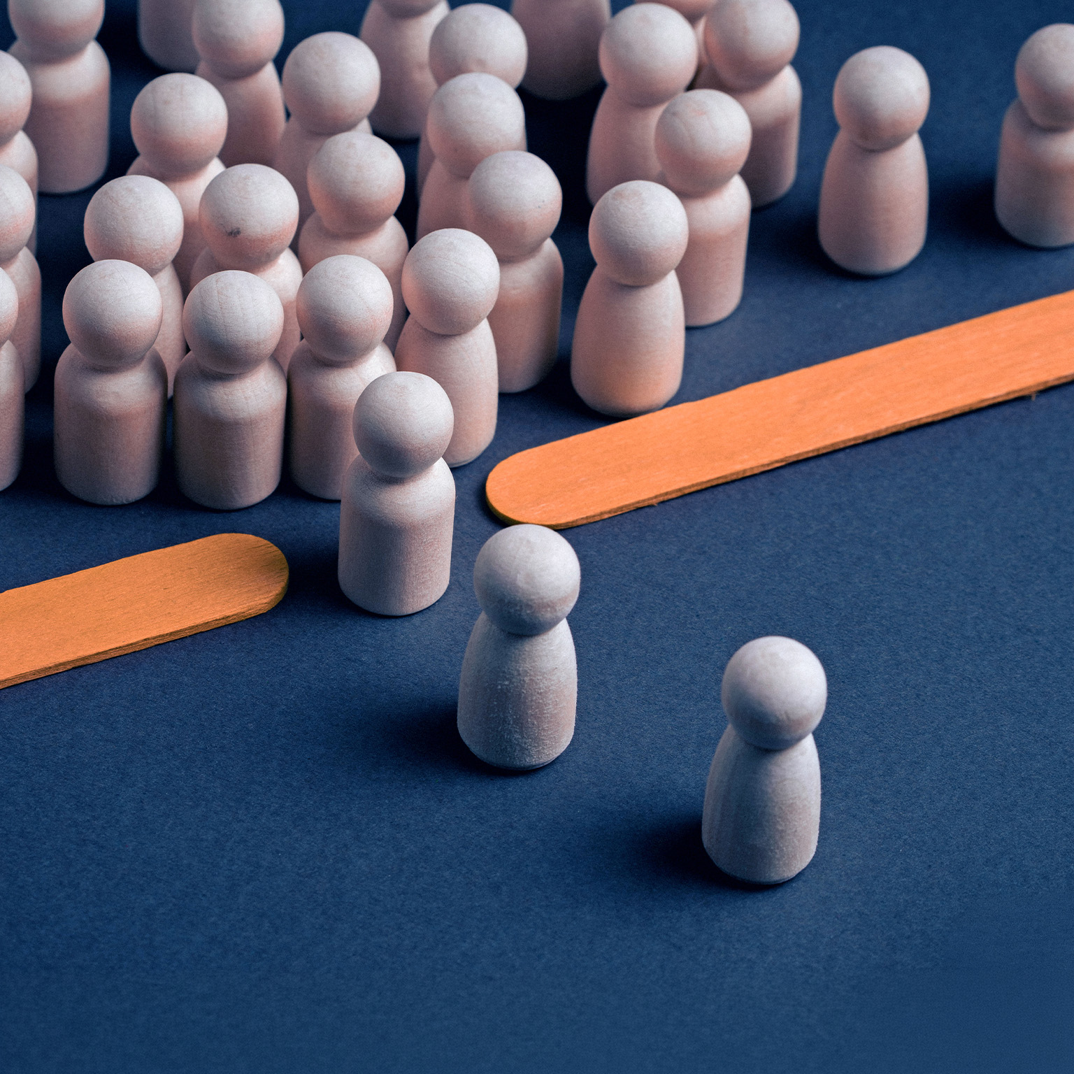 Wooden peg figures arranged on a dark blue surface, with a dense group in the background and several individual figures in the foreground. Two flat orange wooden sticks lie diagonally creating a bottleneck between the two groups.
