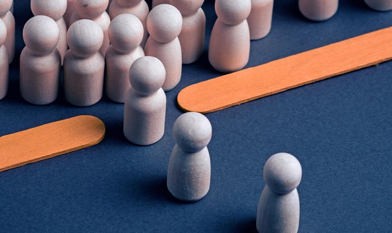 Wooden peg figures arranged on a dark blue surface, with a dense group in the background and several individual figures in the foreground. Two flat orange wooden sticks lie diagonally creating a bottleneck between the two groups.
