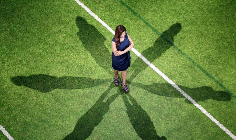 Overhead view of a woman in a dark blue dress standing with her arms crossed on a green sports field, surrounded by six elongated human-shaped shadows. White and dark green field lines cross the grass around her.