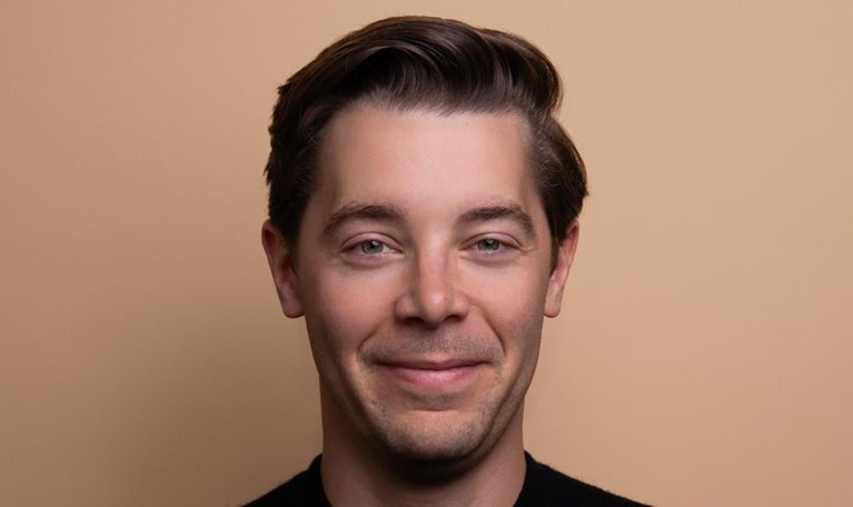 Brad Lightcap with neatly styled dark brown hair looking directly at the camera with a subtle smile. He is wearing a black shirt and is photographed against a plain beige background, creating a simple and professional portrait.