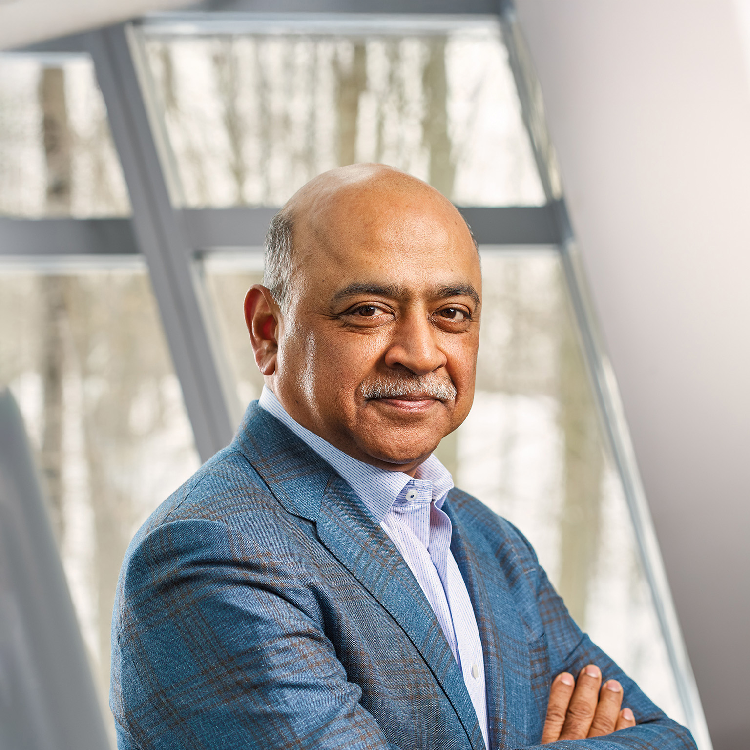 Arvind Krishna with a neatly trimmed mustache, wearing a blue blazer over a light blue collared shirt. He is standing indoors in front of large windows, looking calmly at the camera with a composed, professional expression.