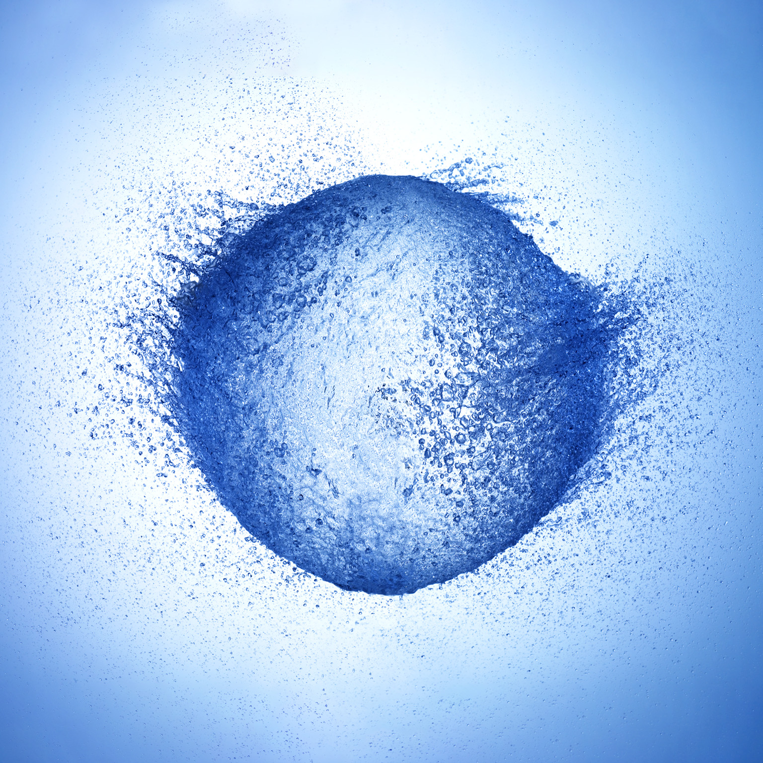 A high-speed photograph of a water balloon bursting. The water is flying outward in all directions, creating a roundish shape with a dynamic and explosive feel.