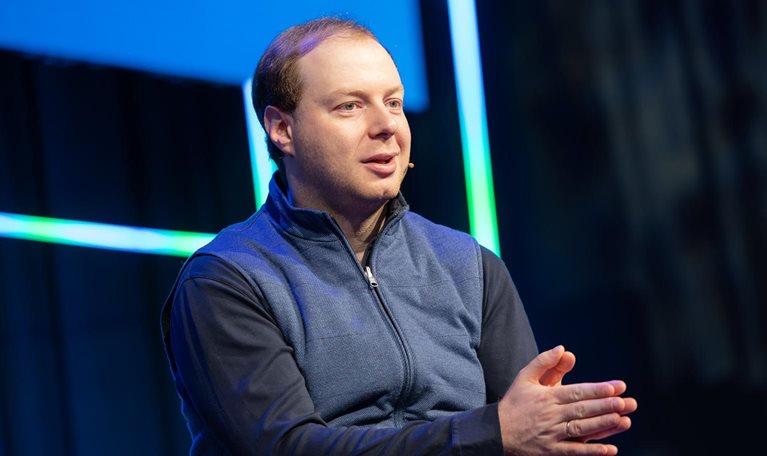 Andrey Khusid speaking on stage with a headset microphone, gesturing with his hands against a dark background accented by blue and green light strips.