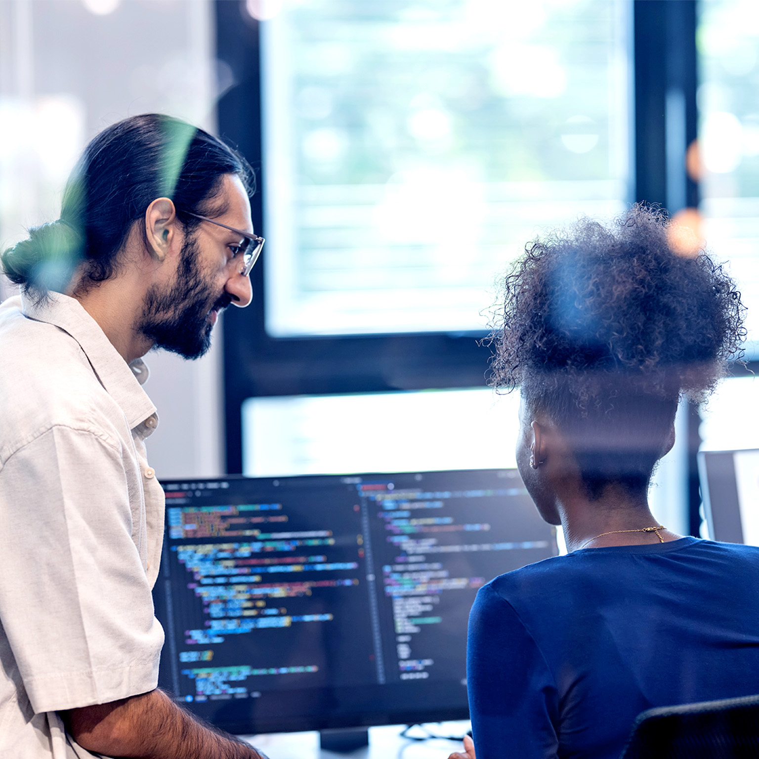 Two software developers collaborate at a workstation, reviewing code displayed on dual monitors in a bright office setting.