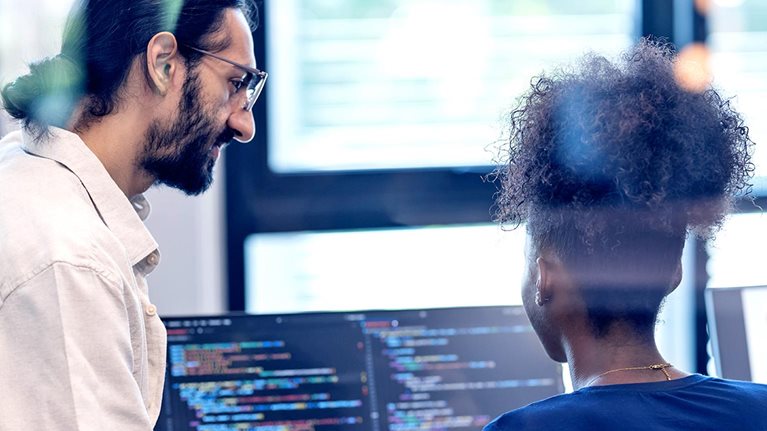 Two software developers collaborate at a workstation, reviewing code displayed on dual monitors in a bright office setting.