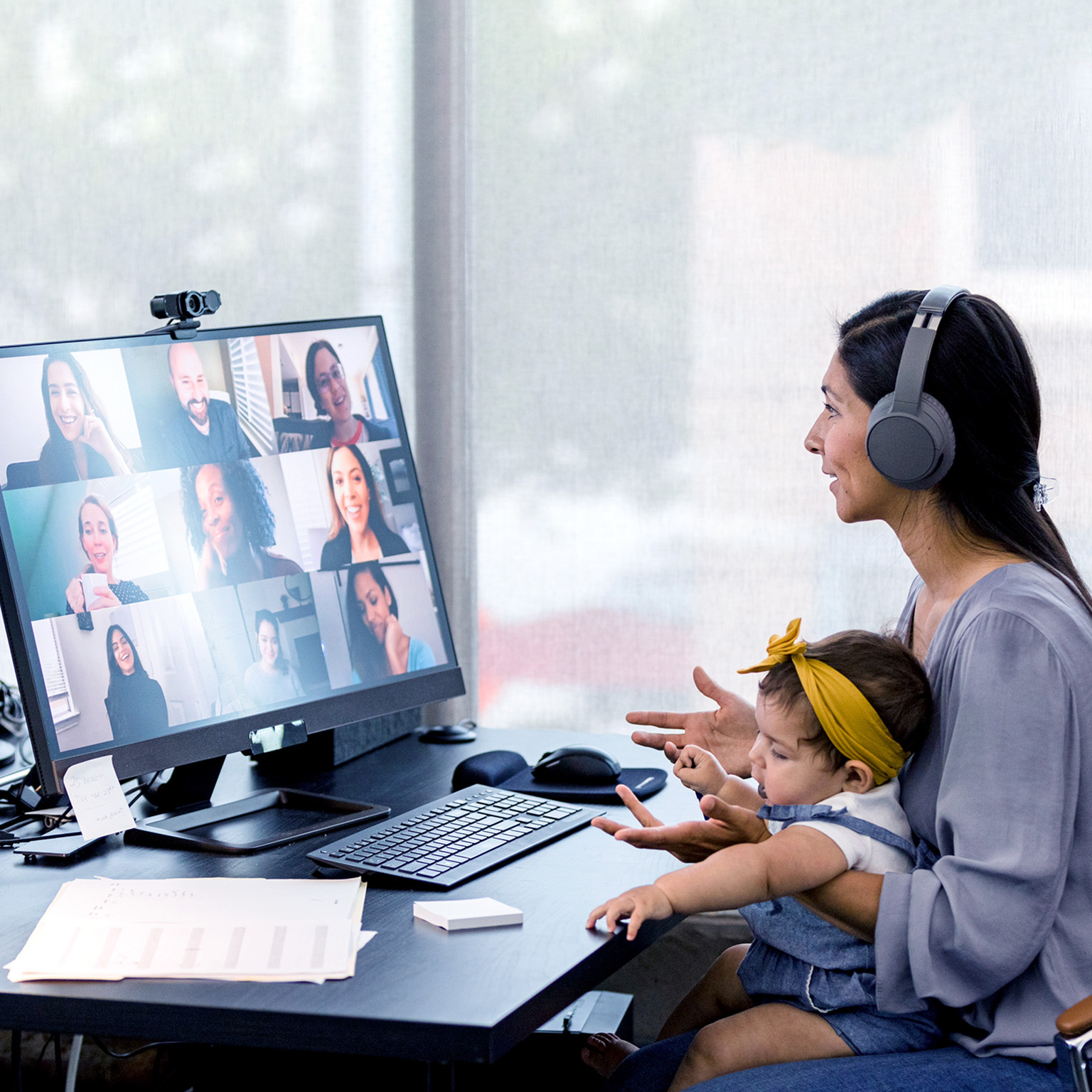 Mother, holding her baby, sits at desk while video conferencing with her co-workers.