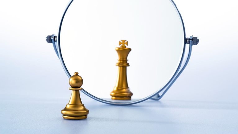 A gold pawn stands in front of a round mirror on a plain surface. In the mirror’s reflection, the pawn appears as a king piece, suggesting a shift in perception or self-image.